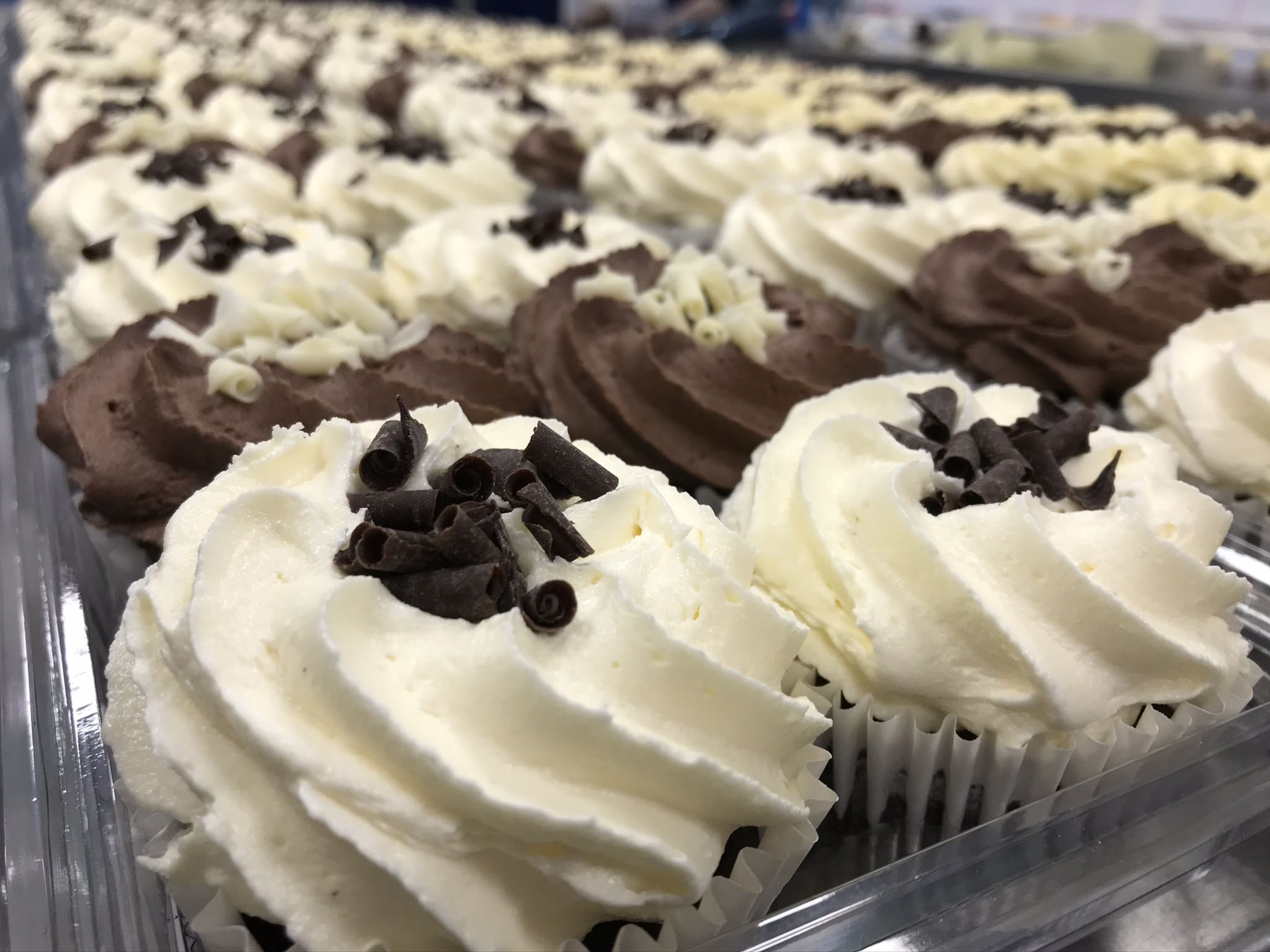 Rows of a dozen mixed cupcakes with white and chocolate swirled frosting are topped with chocolate shavings. The cupcakes are lined up neatly on a tray, highlighting the meticulous presentation and inviting appearance of the personalised assortment.